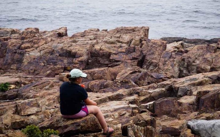 a person sits on a rock along a rocky shoreline while on an outward bound course in maine